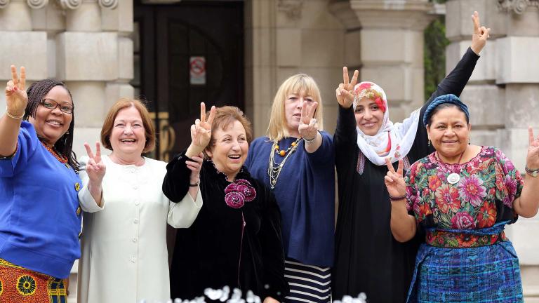 Six femmes debout devant ce qui semble être l’entrée d’un grand édifice de pierre. Elles sourient toutes et lèvent une ou deux mains en faisant le signe de la paix.