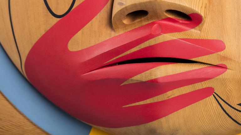 Un plan rapproché d’une boîte en bois, sur laquelle est sculpté un visage dont la bouche est recouverte d’une main peinte en rouge. Visibilité masquée.