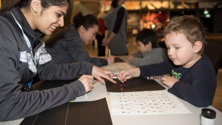 A Museum staff member helping a young child place coloured dots in the shape of braille letters. Visibilité masquée.