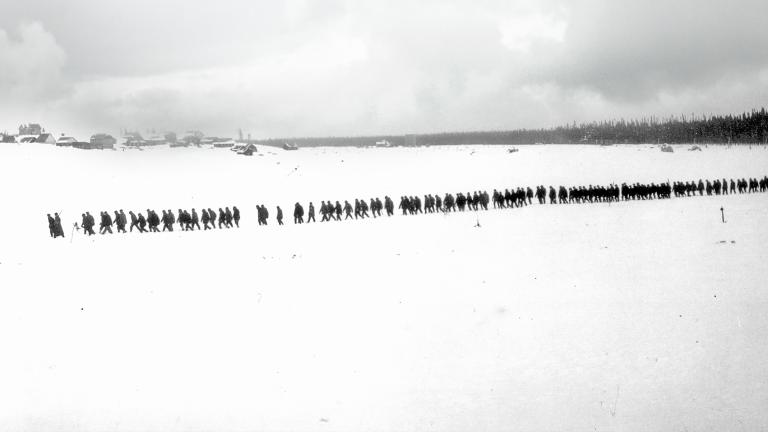 Une photo en noir et blanc d’un paysage plat et enneigé. On voit des arbres et des petits bâtiments au loin. Une longue file de plus de 100 personnes marchent dans la neige. Visibilité masquée.