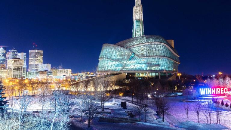 Un bâtiment de verre et d'acier par une soirée enneigée. Devant l'édifice, on voit un signe illuminé qui dit « Winnipeg ». Visibilité masquée.