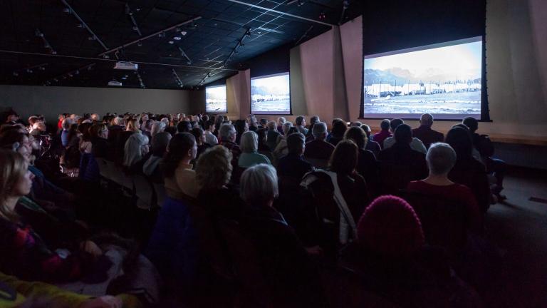 Un auditoire assis regarde trois grands écrans dans une salle dont l'éclairage est tamisé. Visibilité masquée.