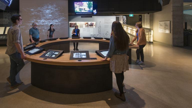Six personnes dans une galerie muséale sont debout à un comptoir circulaire. On voit des écrans vidéo intégrés au comptoir et d’autres suspendus au-dessus des gens. Visibilité masquée.