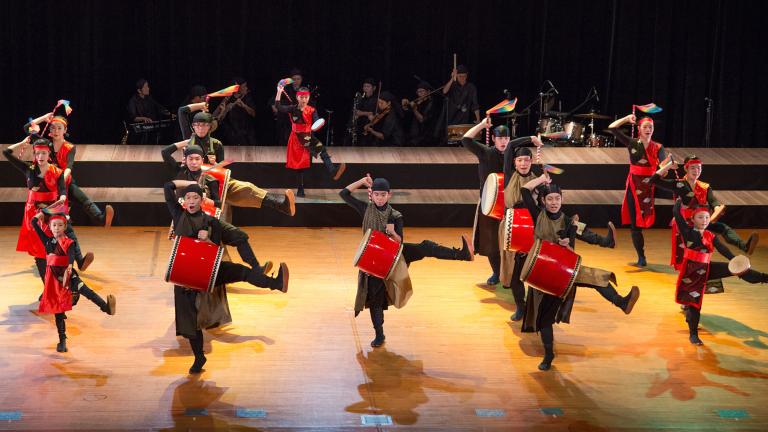 Une troupe de percussionnistes qui danse sur une scène. Les tambours et certains éléments des costumes sont rouge vif. Visibilité masquée.