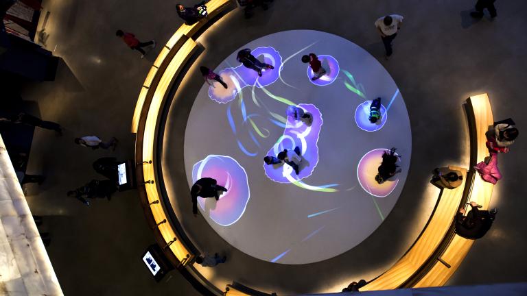 Vue plongeante d’une galerie. On voit environ 9 personnes à l’intérieur d’un grand cercle sur le plancher. Des lumières colorées créent des cercles lumineux autour de leurs pieds. D’autres personnes sont assises sur des bancs autour du cercle. Visibilité masquée.