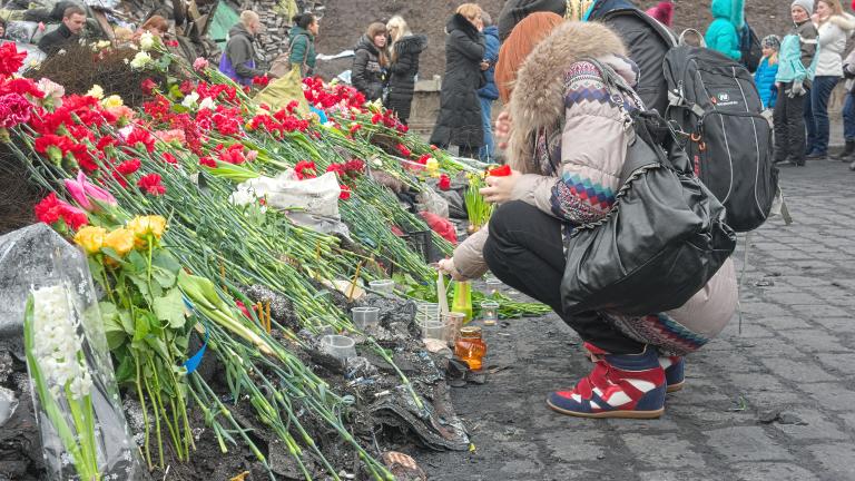 Deux personnes déposant des fleurs rouges à un mémorial. En arrière-plan, une douzaine d’autres personnes se tiennent devant des débris, comme des roches, des feuilles de métal et des barrières de ciment peintes avec des drapeaux ukrainiens bleu et jaune et des lettres. Visibilité masquée.