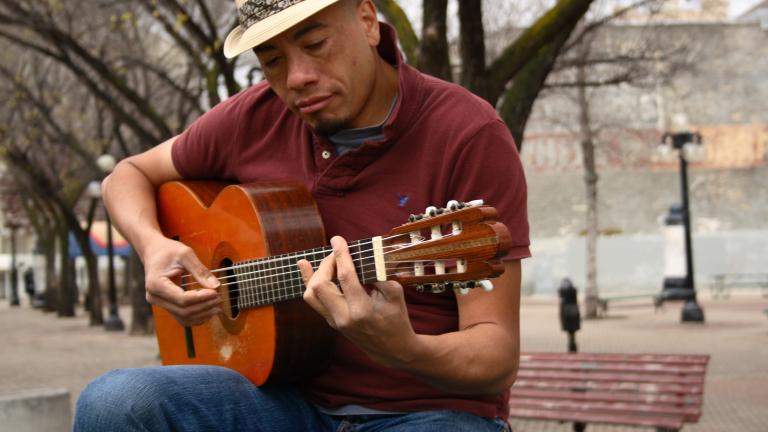A man wearing a fedora plays a guitar while sitting outside on a bench. There are trees in the background. Visibilité masquée.