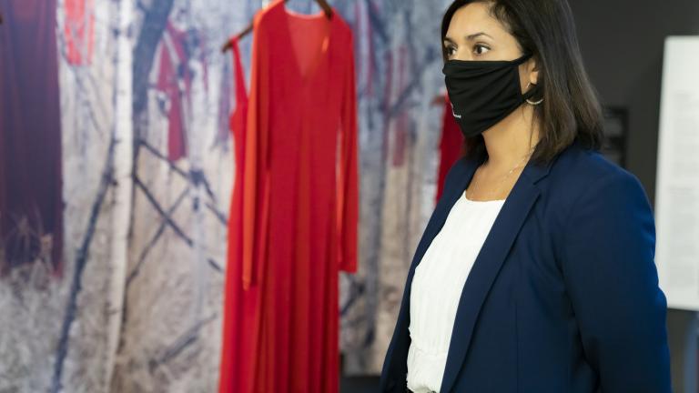 Une femme portant un masque noir se tient devant une exposition du Musée composée de robes rouges suspendues devant un fond de forêt. Visibilité masquée.