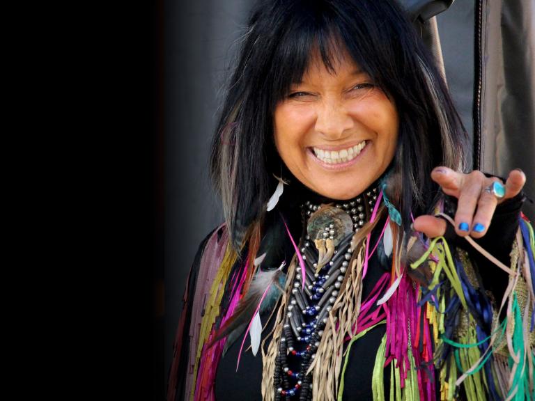 Une femme autochtone aux cheveux noirs sourit et porte un habit élégant décoré de perles et de nombreux rubans colorés. Visibilité masquée.