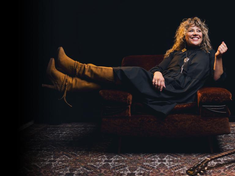 Une femme très heureuse, avec des cheveux blonds bouclés et une guitare à ses pieds, est assise les pieds en l'air sur un canapé. Visibilité masquée.