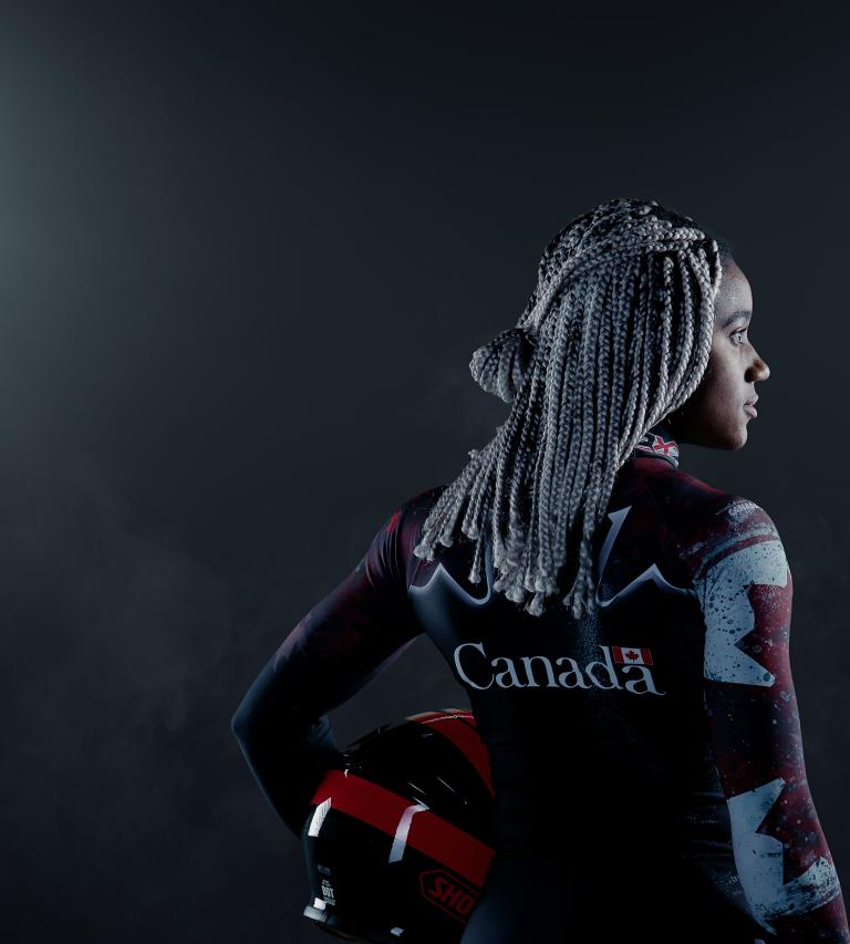 Une femme noire se tient dans un endroit sombre entre deux grands projecteurs. Elle tient un casque et porte une combinaison de bobsleigh canadienne. Visibilité masquée.