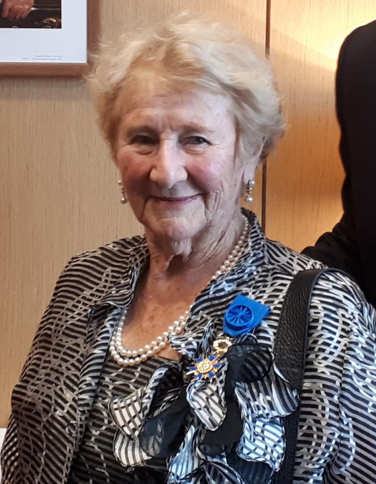 Une femme âgée aux cheveux courts et gris sourit doucement à la caméra. Elle porte des boucles d’oreilles en perles, un collier de perles et un chemisier à motifs noir et blanc. Elle se tient debout, baignée d’une lumière chaleureuse, devant un mur lambrissé. Visibilité masquée.
