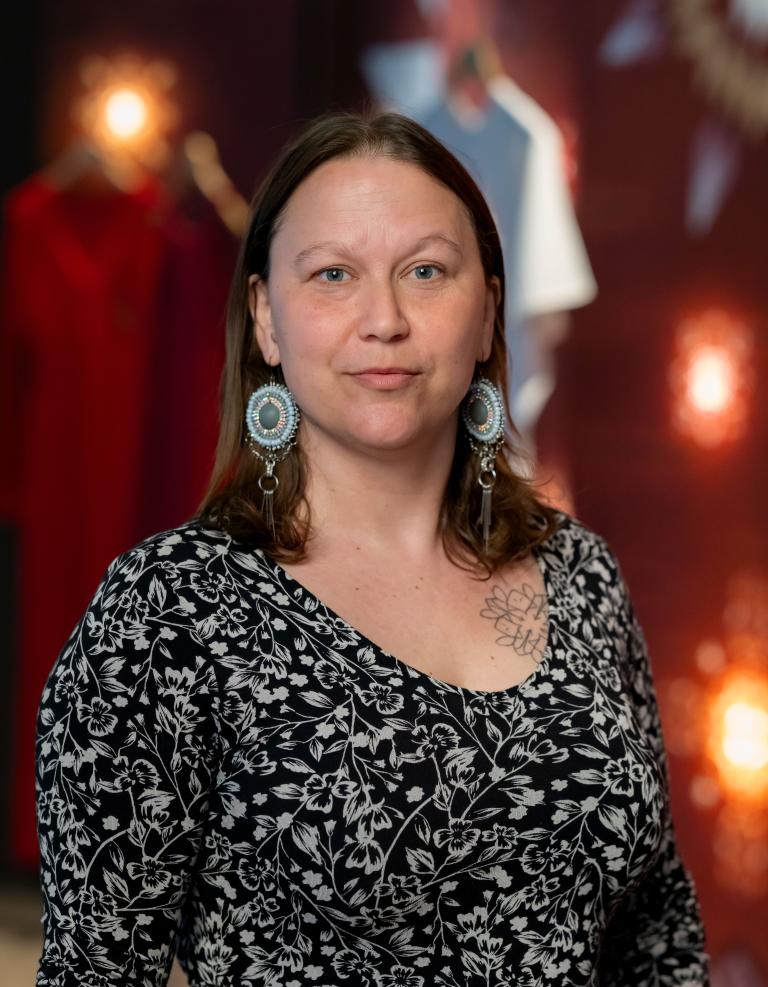 Une femme se tient fièrement devant des robes rouges en arrière-plan. Elle porte une robe à fleurs noires et blanches et des boucles d'oreilles circulaires bleu clair. Visibilité masquée.