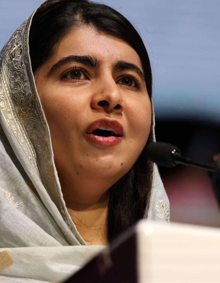 Malala Yousafza, une jeune femme portant un hidjab de couleur claire, parle dans un microphone. Visibilité masquée.