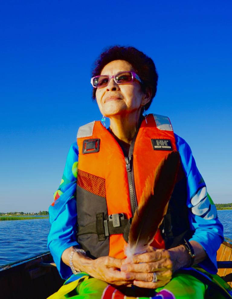 Une femme autochtone se trouve dans un canot sur un lac avec un gilet de sauvetage orange. Elle semble sereine. Visibilité masquée.
