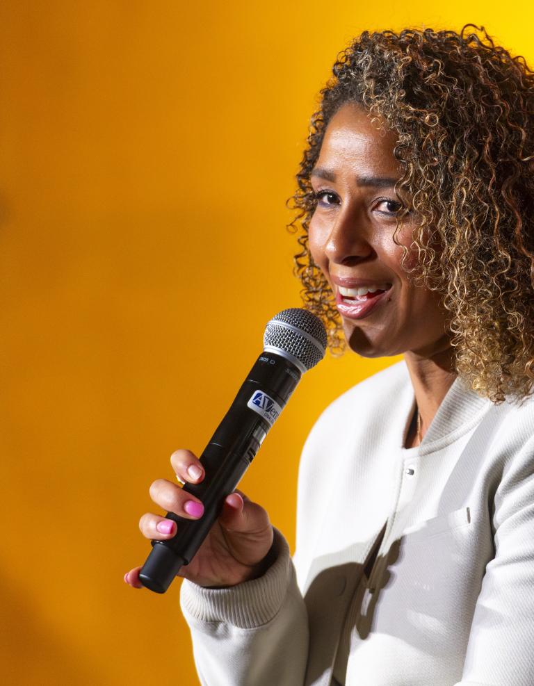 Une femme aux cheveux bouclés parle dans un micro à main lors d’une table ronde, souriant chaleureusement devant un fond jaune vif. Visibilité masquée.