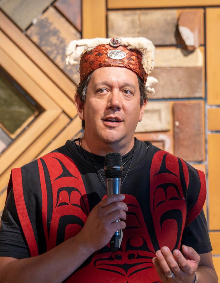 Un homme debout, micro à la main, s’adresse à une foule vêtue de costumes traditionnels autochtones devant une installation artistique en panneaux de bois. Visibilité masquée.
