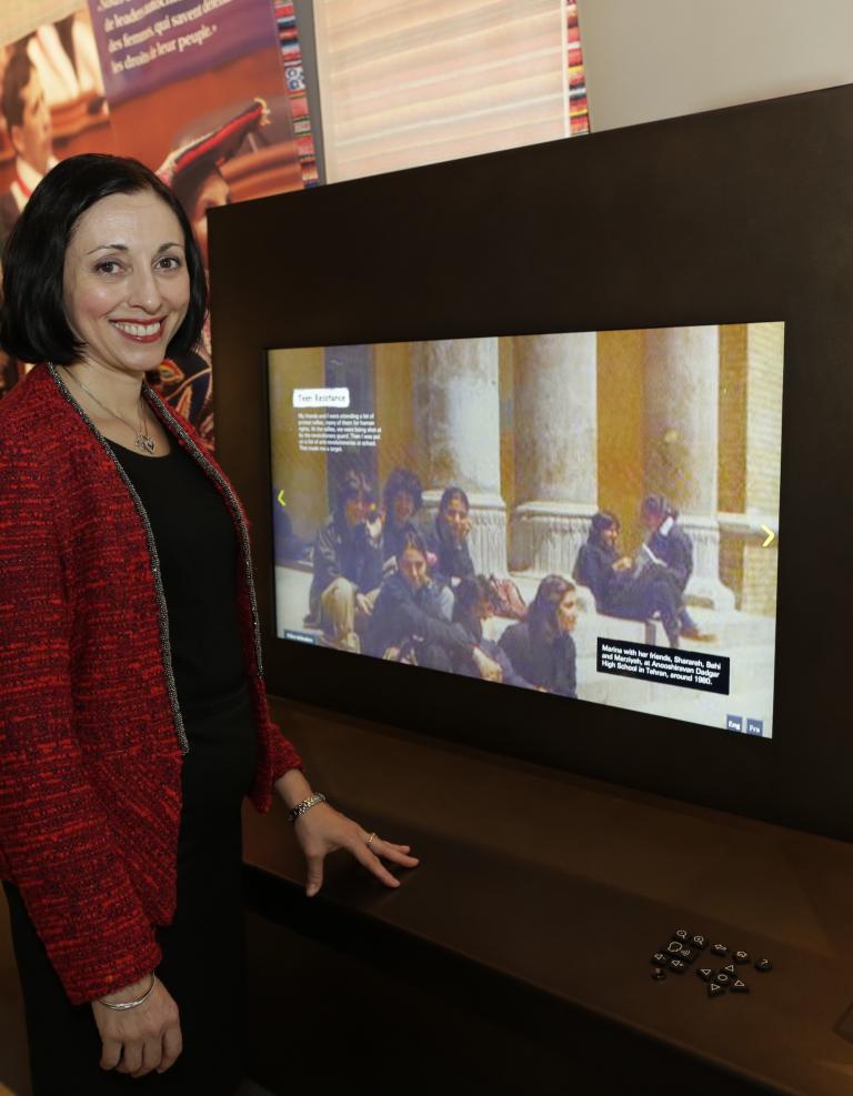 Marina Nemat sourit alors qu’elle se tient debout à côté d’un écran numérique de grande taille. Elle porte une veste rouge et regarde directement vers l’appareil photo. Sur l’écran, on peut voir une image montrant des personnes assises devant d’énormes colonnes de pierre. 