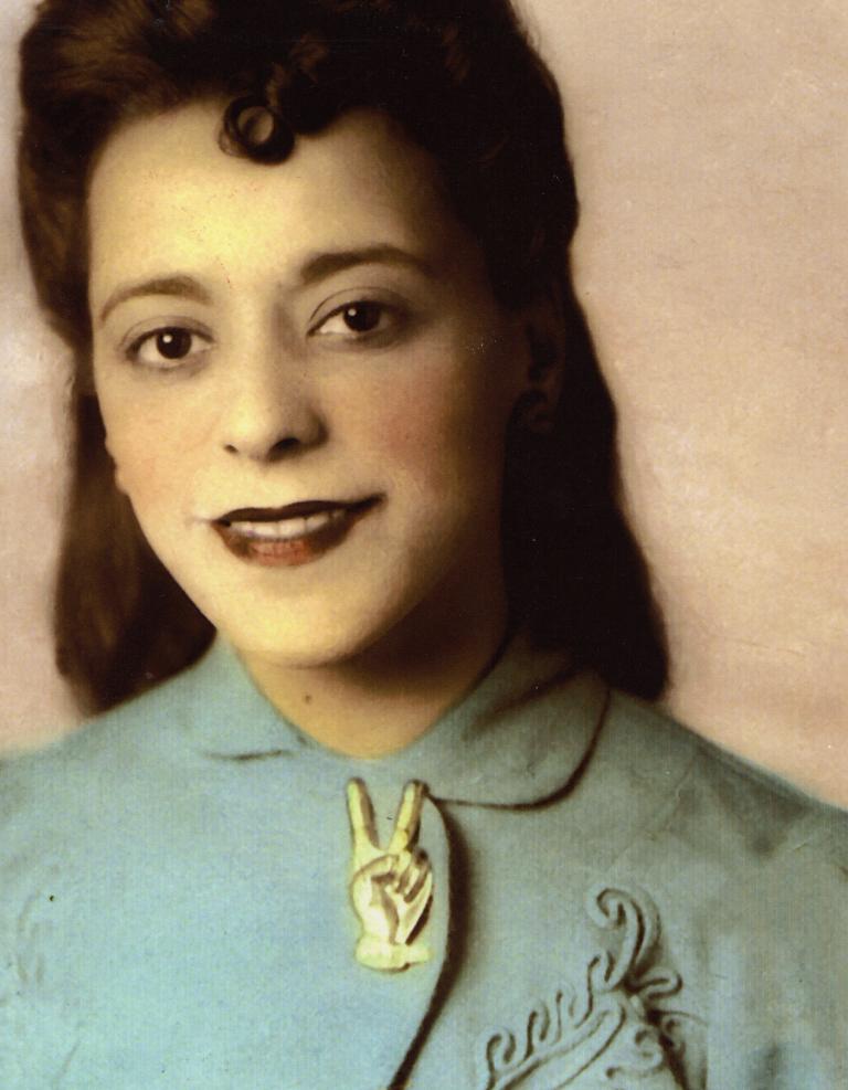 Portrait en buste d’une Viola Desmond souriante. Elle porte une veste bleu pâle à motifs brodés. La veste est fermée au col par une broche en forme de main avec deux doigts écartés pour former le V de la victoire.