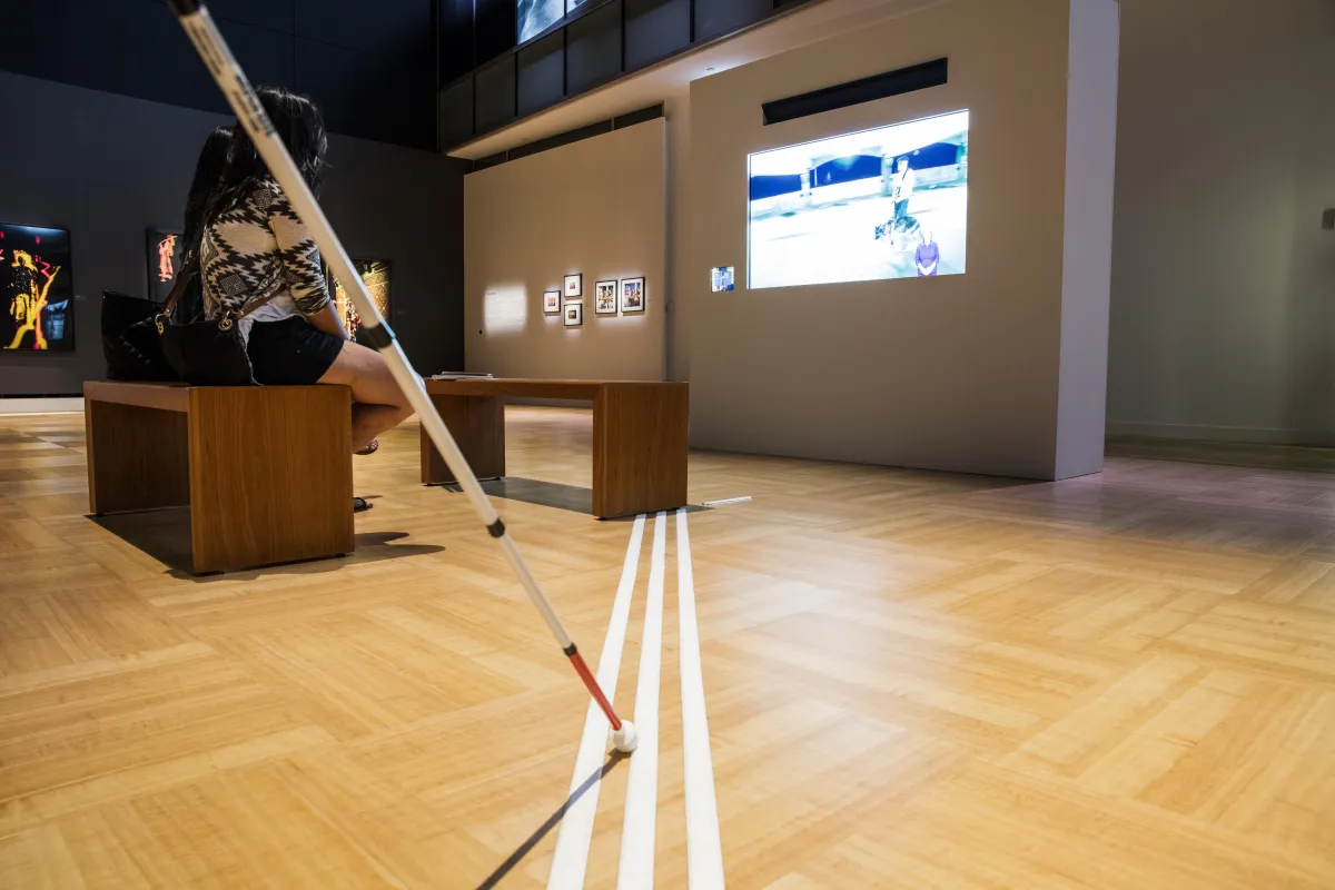 Au premier plan, la canne blanche d'une personne malvoyante touche trois bandes tactiles disposées sur le sol d'une galerie du Musée. Ces bandes mènent à des bancs, où deux personnes regardent un film sur grand écran.