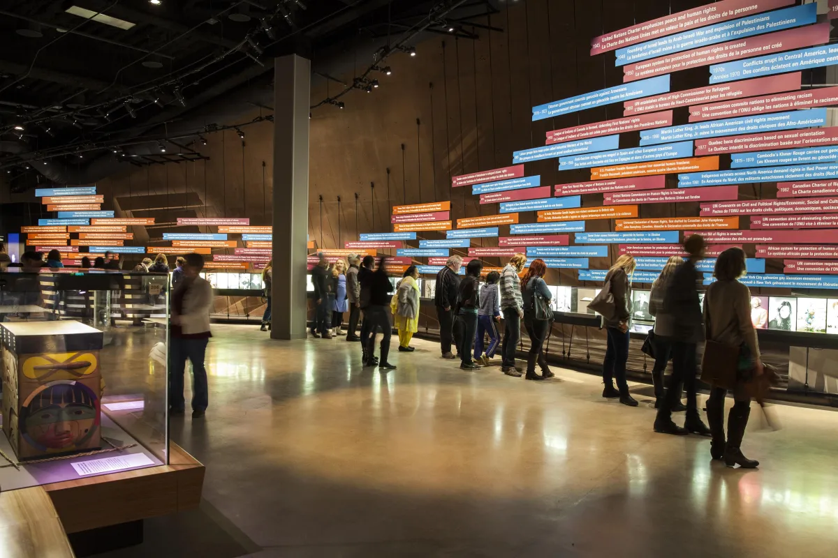 Une grande foule dans une galerie du Musée. La plupart des gens regardent une exposition sur le mur de la galerie. L'exposition comprend une série d'images et de longs panneaux rectangulaires de texte, placés horizontalement.
