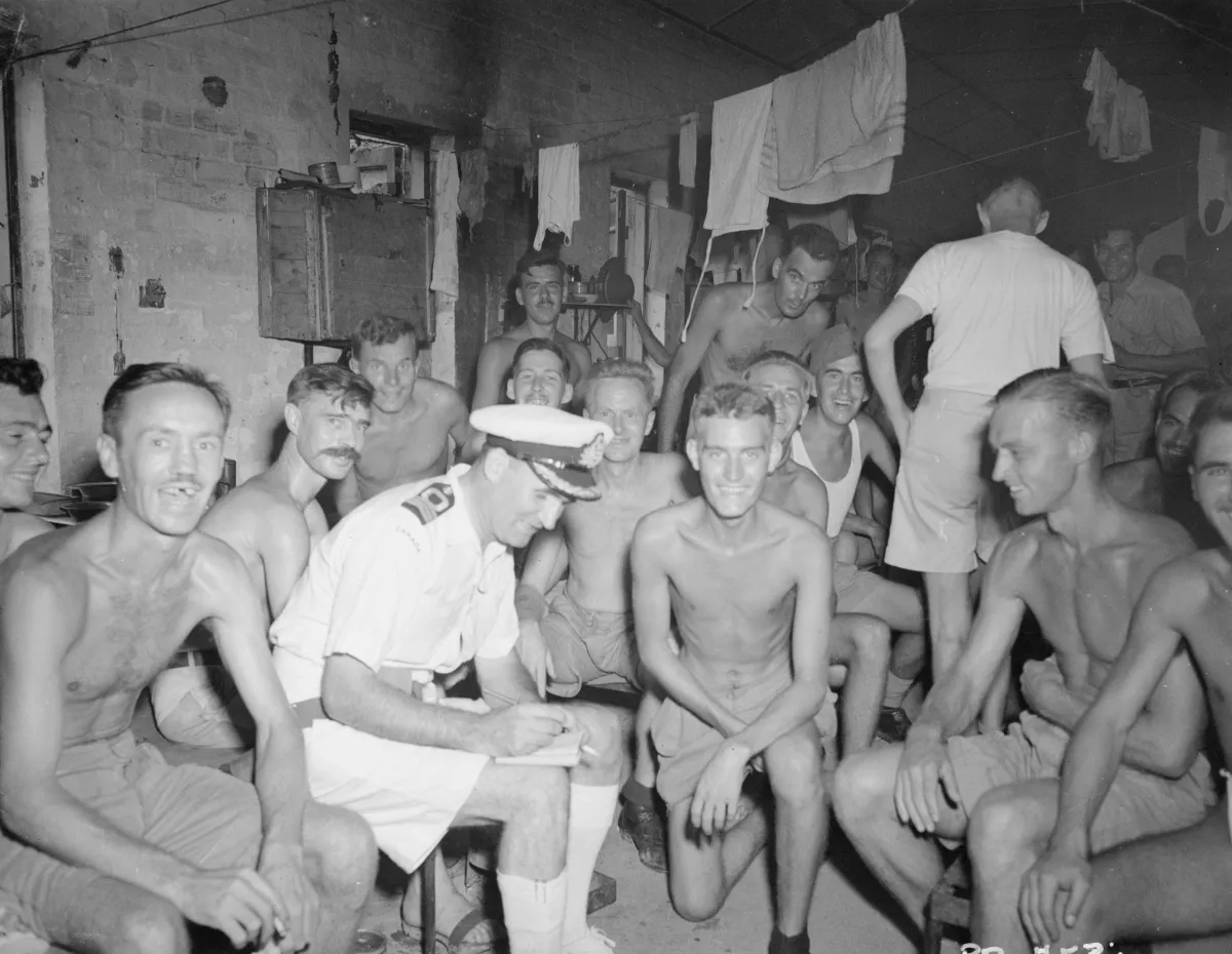 Photo noir et blanc où l’on voit plus d’une douzaine d’hommes rassemblés dans une petite salle sombre. Plusieurs sourient à la caméra. La majorité sont torse nu et émaciés.
