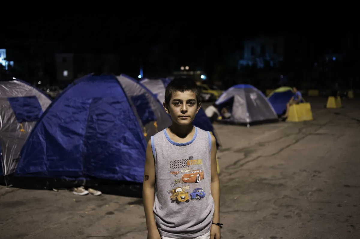 Un adolescent debout devant des tentes installées dans un terrain de stationnement. Il fait nuit.