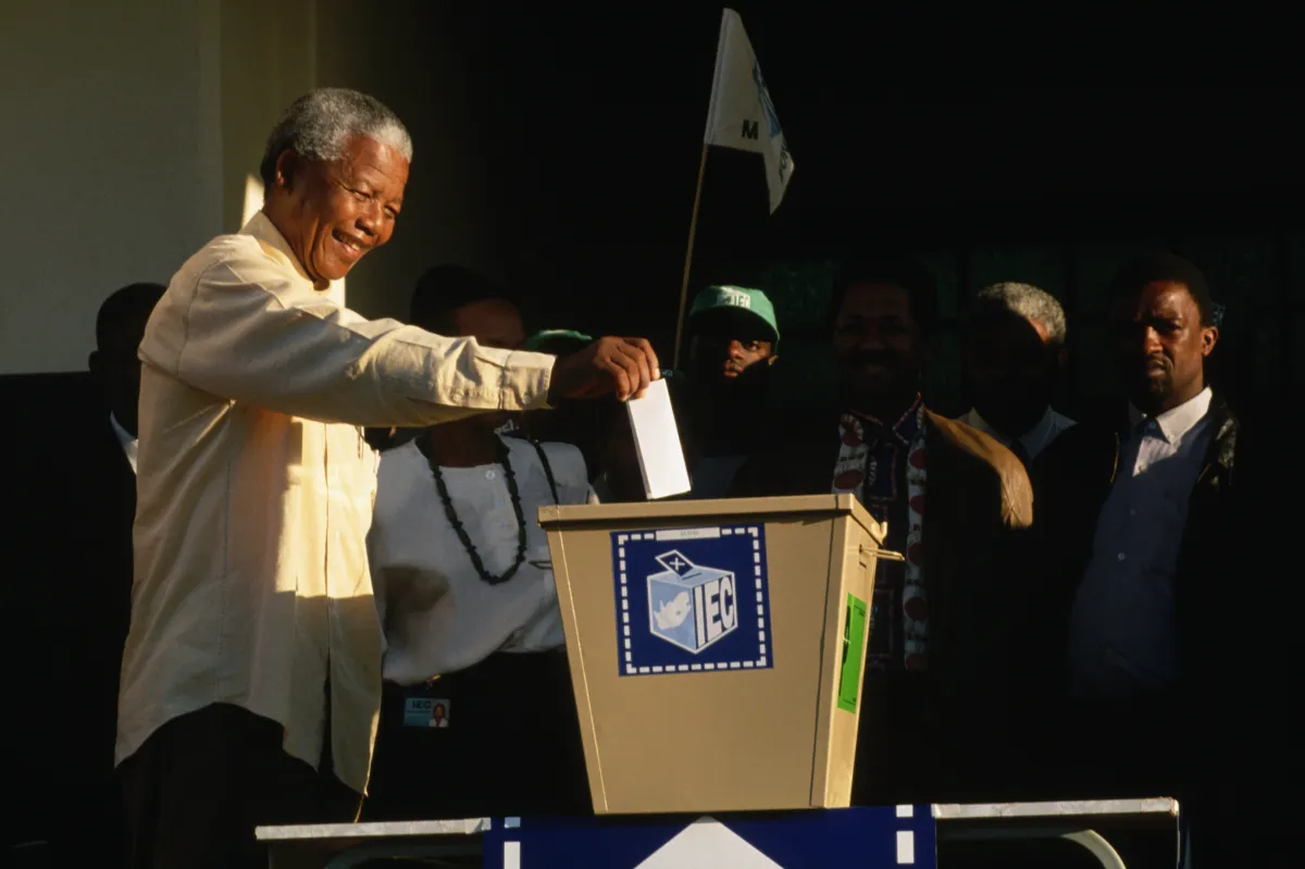  Nelson Mandela étendant son bras droit pour déposer un papier plié dans une boîte de scrutin. Quelques personnes sont debout autour de lui.