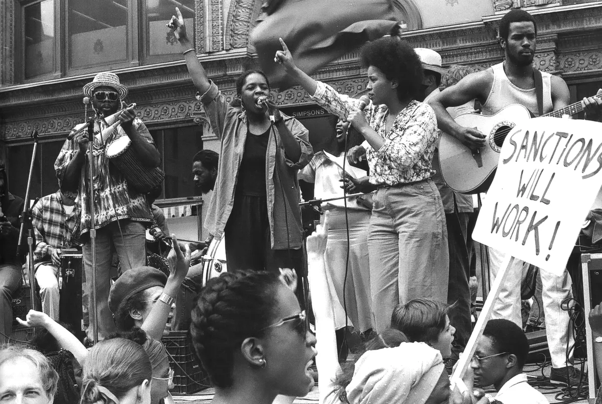 Des hommes et des femmes musiciens sur une scène. Les deux chanteuses au centre ont le bras droit levé. Dans la foule qui les regardent, on voit des pancartes de protestation. 