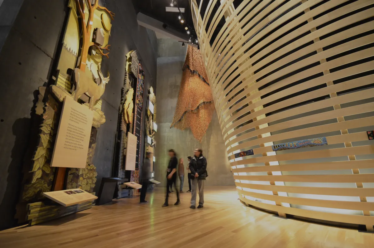 Quelques personnes dans une galerie du Musée explorent de grands panneaux de bois décorés de figures d’animaux et d’une œuvre perlée métisse. À droite, on voit des lattes de bois courbées placées horizontalement.