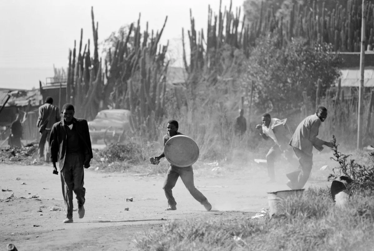 Photo en noir et blanc de quelques hommes noirs dans une environnement extérieur poussiéreux. L’un d’eux tient un couvercle de poubelle en guise de bouclier.