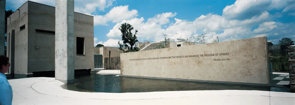Un vue du Apartheid Museum. À l’avant-plan, on voit un plan d’eau. Dans l’eau se dresse un mur sur lequel on peut lire en anglais une citation de Nelson Mandela qui dit : « To be free is not merely to cast off one’s chains, but to live in a way that respects and enhances the freedom of others. »