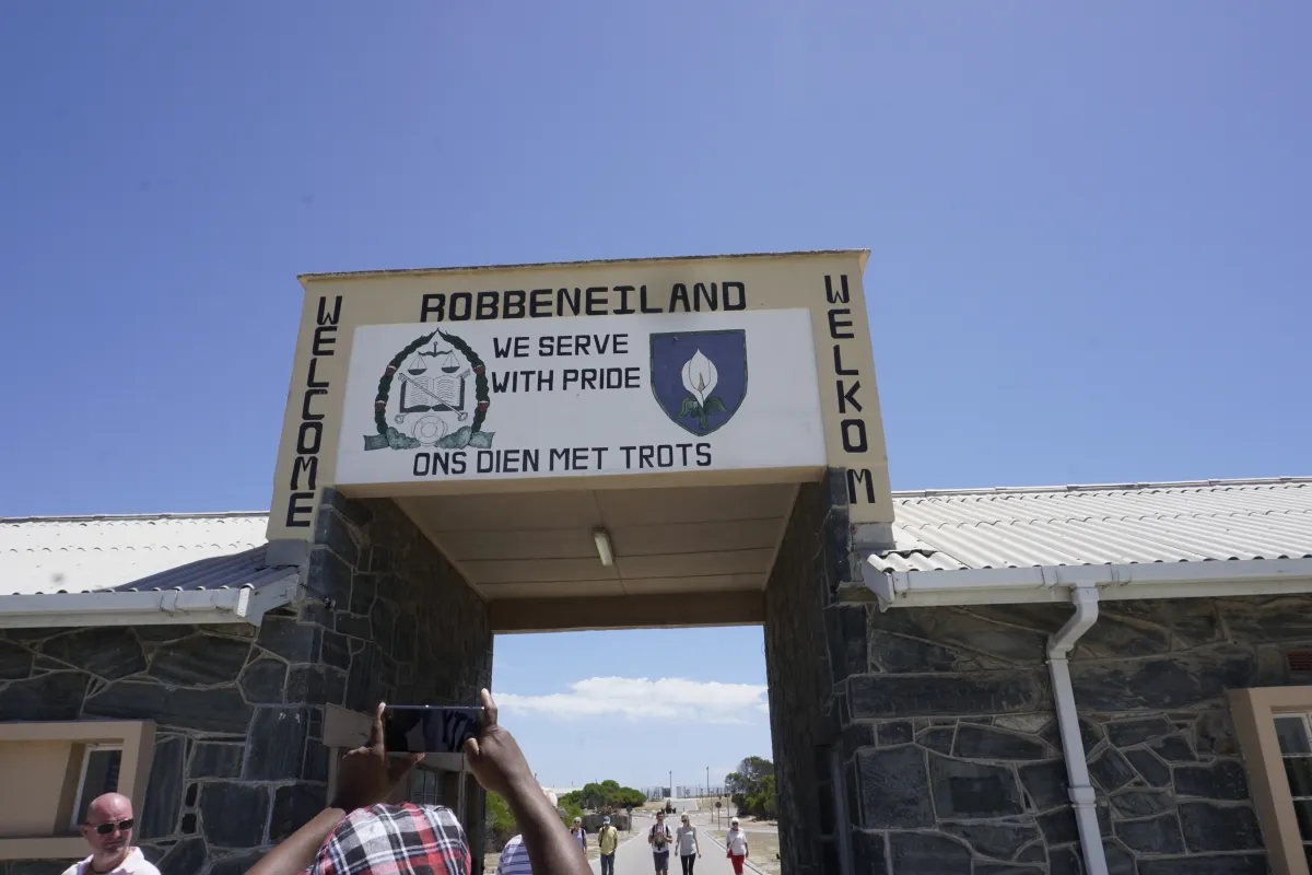Une ouverture dans un bâtiment et un grand panneau au-dessus de l’ouverture. Sur panneau, on peut lire en anglais et en afrikaans « Welcome » et « We serve with pride ». Au haut du signe, on peut lire « Robbeniland ».