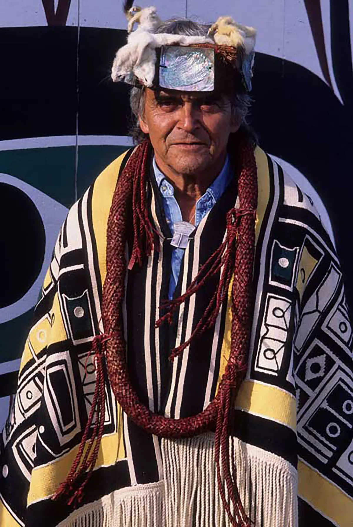 Bill Cranmer, un homme plus âgé aux cheveux gris, se tient debout vers l’appareil photo. Il porte une couverture à franges et aux motifs géométriques sur ses épaules. Il porte aussi un chapeau de fourrure orné d’une pièce centrale de forme carrée en métal brillant et un gros collier fait de corde épaisse.