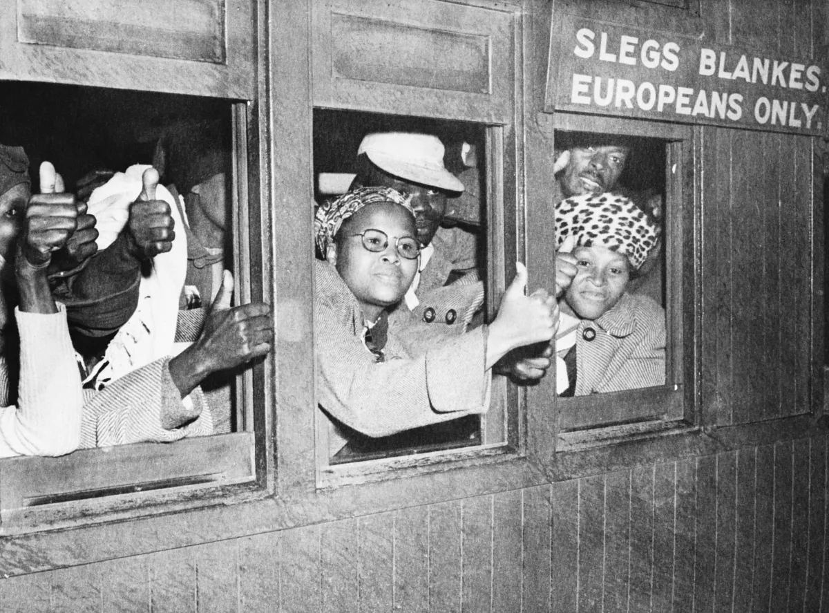 Une image en noir et blanc d’un groupe de femmes et d’hommes noirs à l’intérieur d’un wagon qui regardent à l’extérieur par les fenêtres. Plusieurs ont la main levée et tiennent le pouce en l’air. Au-dessus d’une fenêtre, il est écrit que le wagon est réservé à la population européenne.