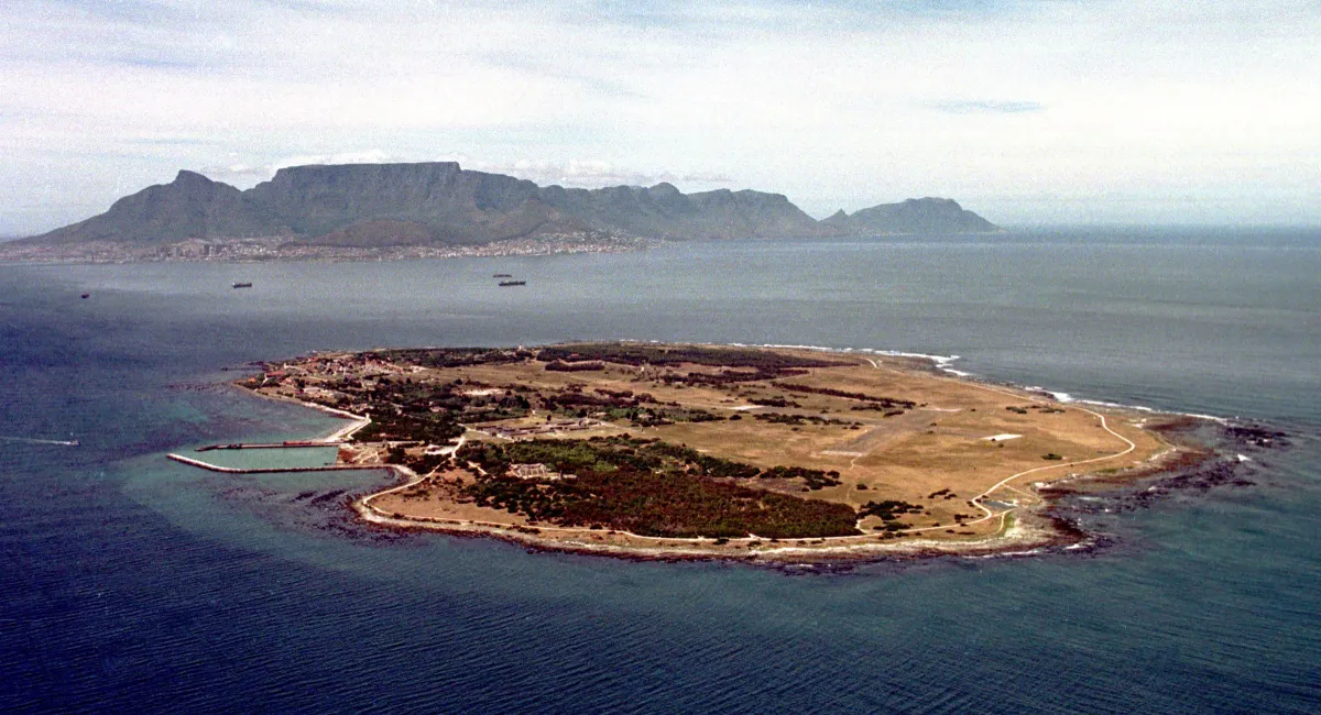 Vue aérienne de l’île Robben. L’île est plus ou moins de forme ovale et le terrain est très plat et couvert presqu’à moitié d’arbres. On aperçoit un terrain d’aviation sur le côté droit de l’île. Au loin, derrière l’île, on voit le continent, y compris les édifices d’une ville. Au-delà de la ville se dressent de grandes montagnes.
