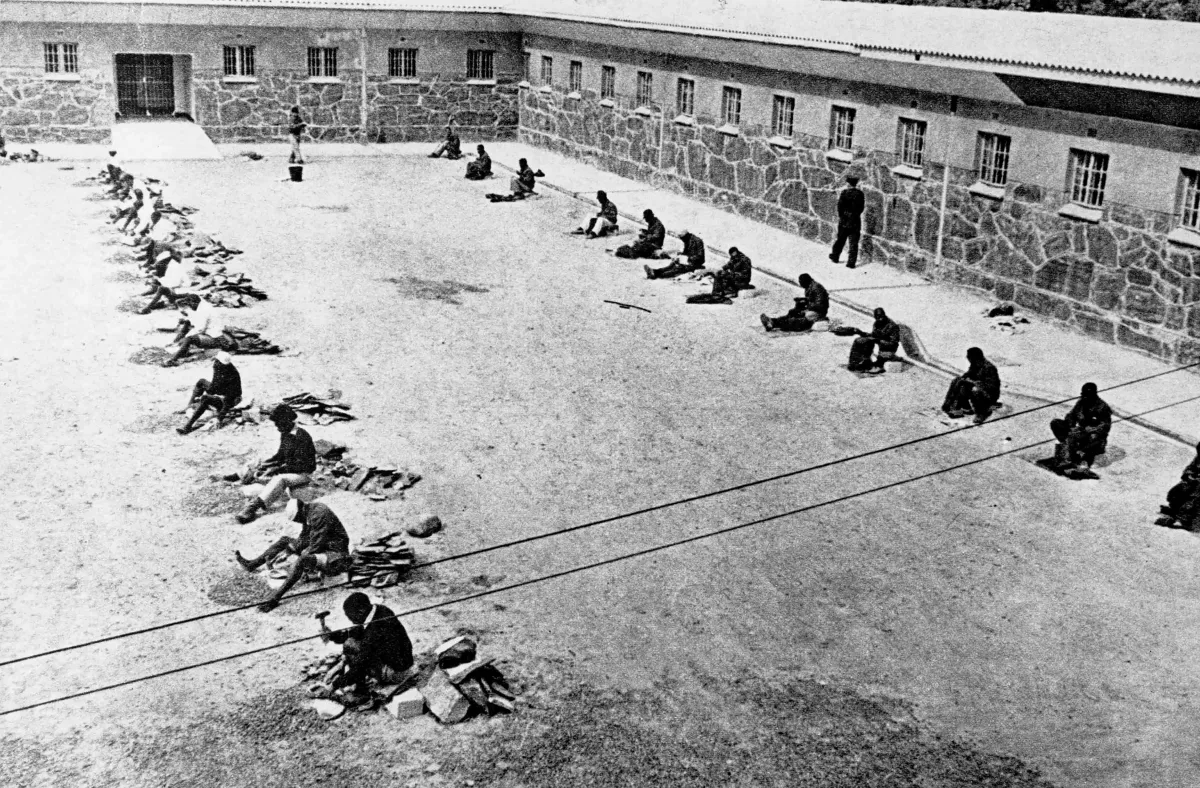 Une image en noir et blanc où l’on voit deux rangées d’hommes dans une cour. Ils sont assis et brisent des roches à l’aide de petits marteaux.