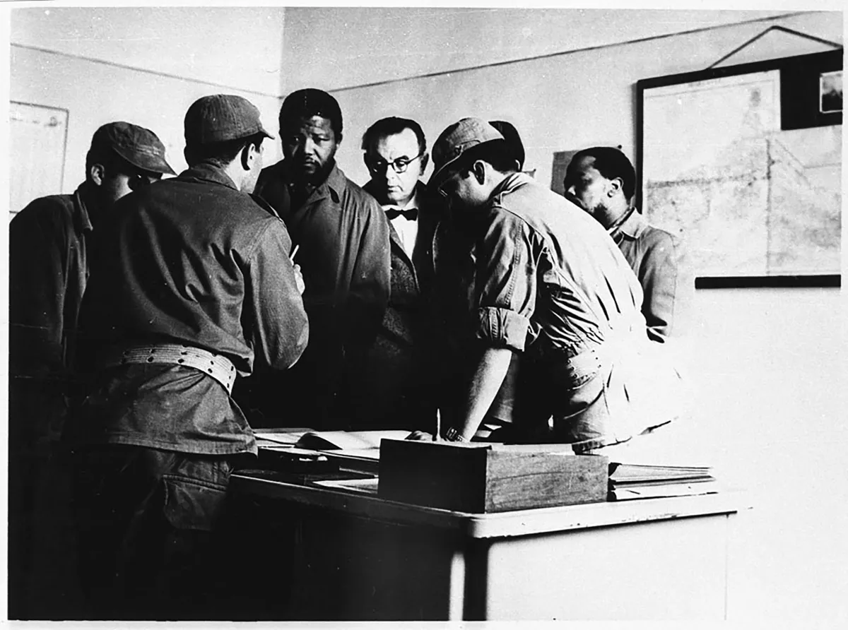 Une image en noir et blanc de Nelson Mandela debout devant un bureau. Il est entouré de cinq hommes, certains portant une tenue militaire. Un homme, le dos tourné à l’objectif, semble parler à Mandela, et les autres regardent quelque chose sur le bureau. 