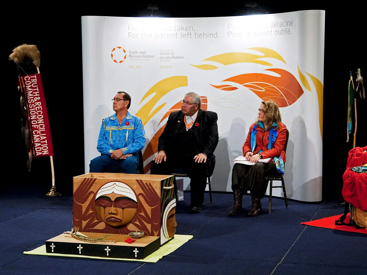 Deux hommes et une femme assis devant un arrière-plan blanc décoré de flammes stylisées. À leur droite, on voit une bannière verticale sur laquelle on peut lire « Truth & Reconciliation Commission of Canada ». et devant eux se trouve une boîte rectangulaire en bois sculpté. Sur le côté qui fait face à l'objectif, on voit un visage rond qui pleure.