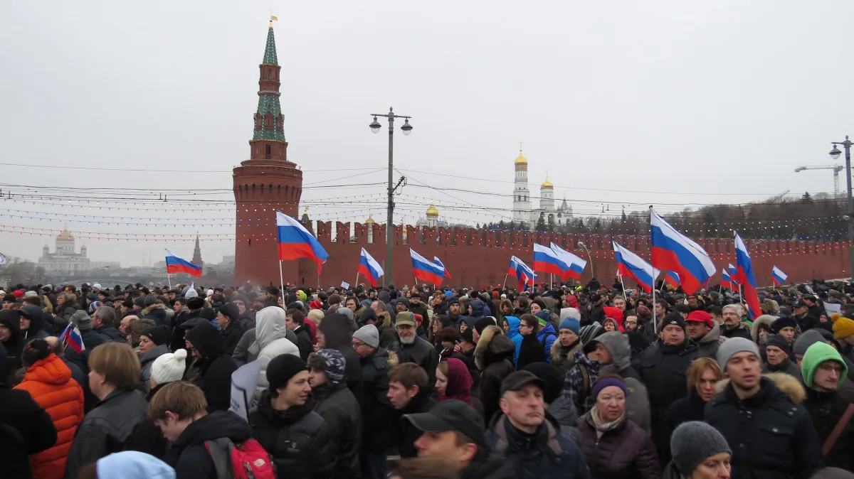 Une foule marche; plusieurs personnes portent le drapeau aux rayures rouge, blanche et bleue de la Russie. Derrière, on voit un mur de béton rouge qui se termine en flèche.