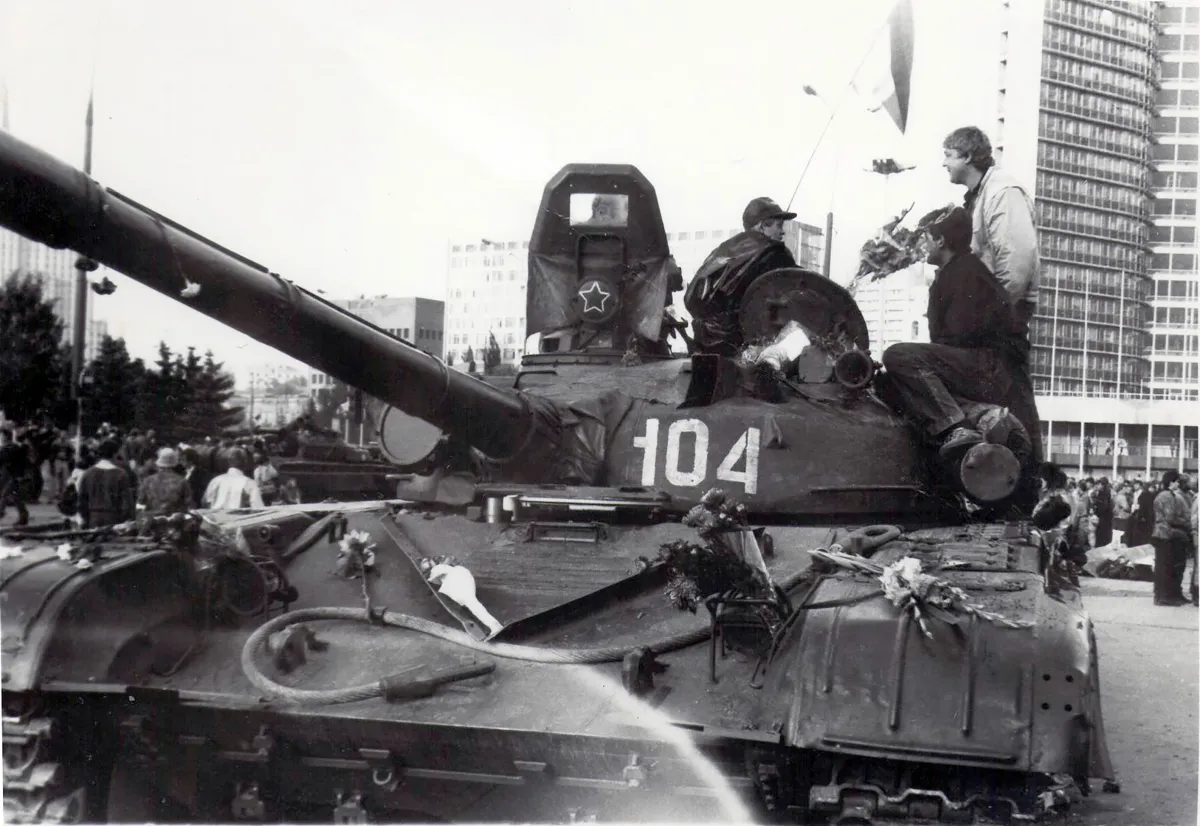 Deux hommes debout devant un char militaire stationné. Il y a des bouquets de fleurs sur le char. Derrière, on voit des gens déambuler devant un haut édifice. La photo est en noir et blanc.