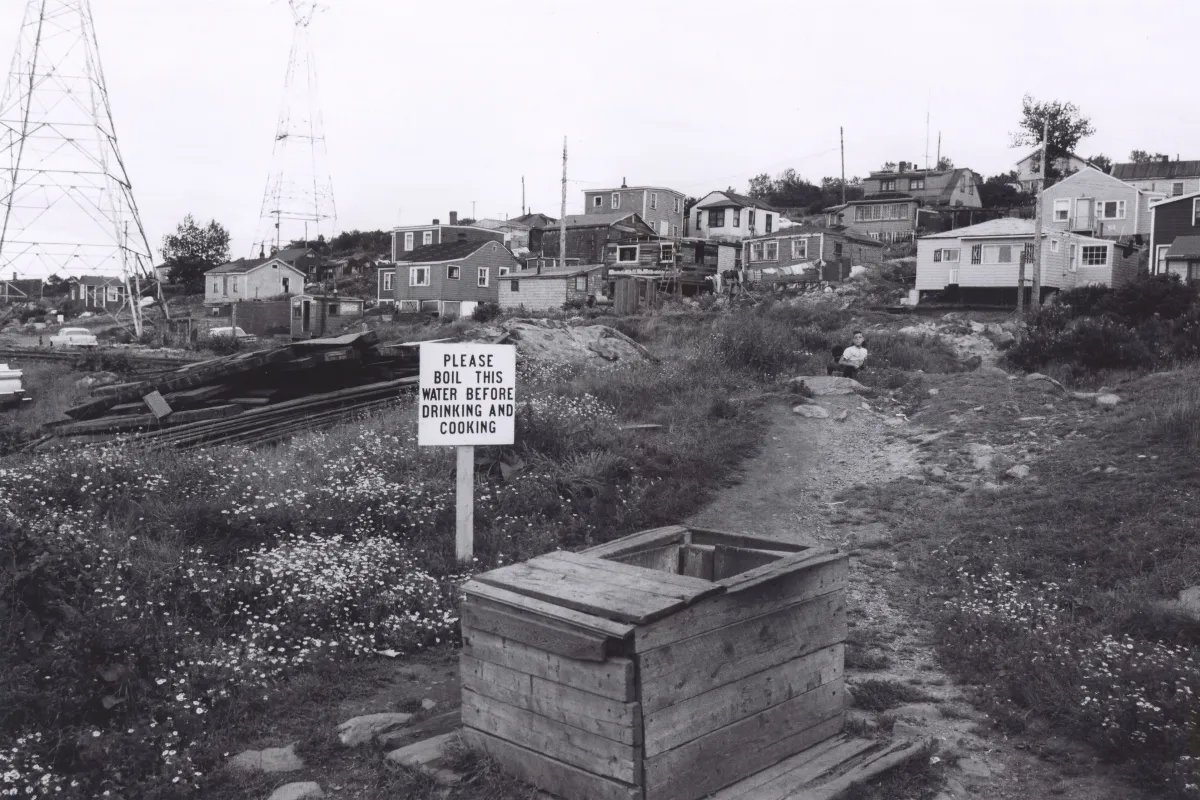 Une image en noir et blanc d’environ 15 maisons sur une colline. À l’avant-plan, on voit un puits et un signe à côté qui dit, en anglais : « Veuillez faire bouillir cette eau avant de la boire ou de l’utiliser pour la cuisson. »