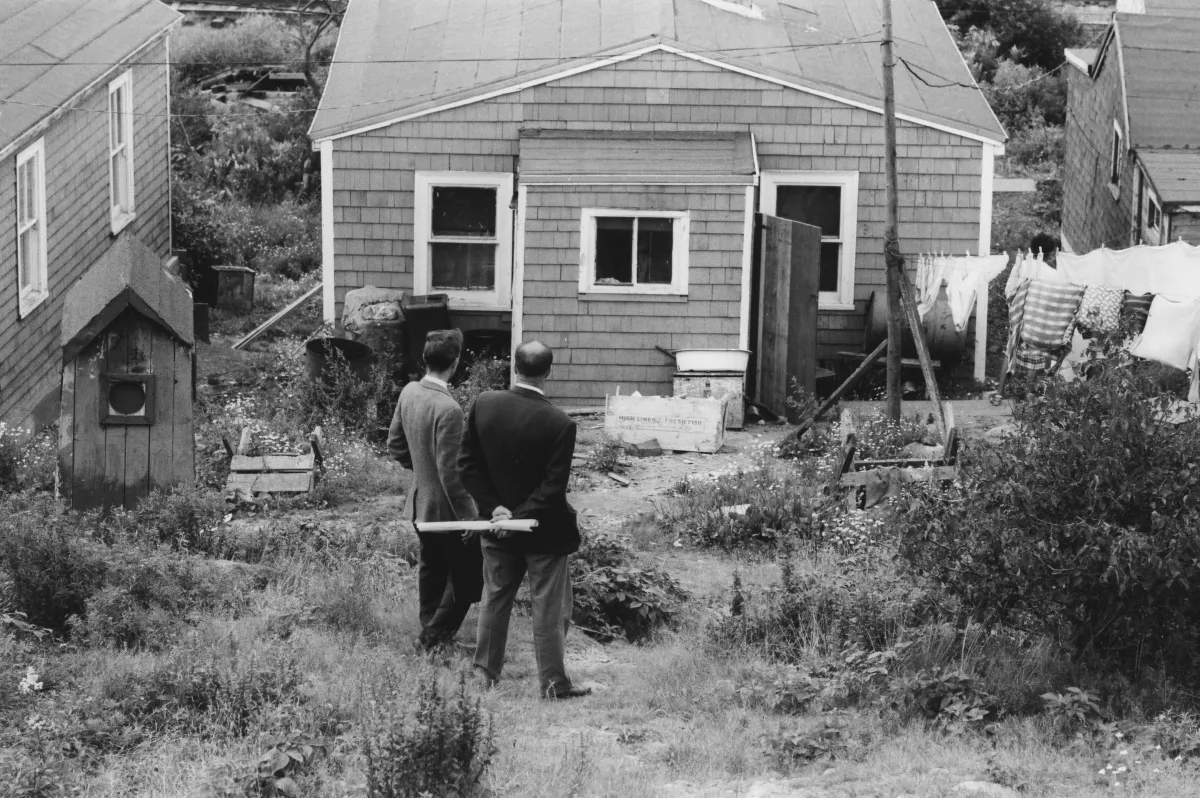 Une image en noir et blanc de deux hommes en complet debout devant une maison en bois. Ils font dos à l’objectif.