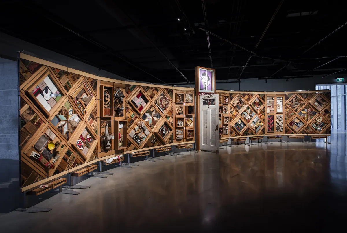 Une grande installation artistique horizontale à armature en bois composé de multiples losanges et rectangles sur lesquels sont fixés de nombreux objets. Une porte est entrouverte en plein centre de l’œuvre.