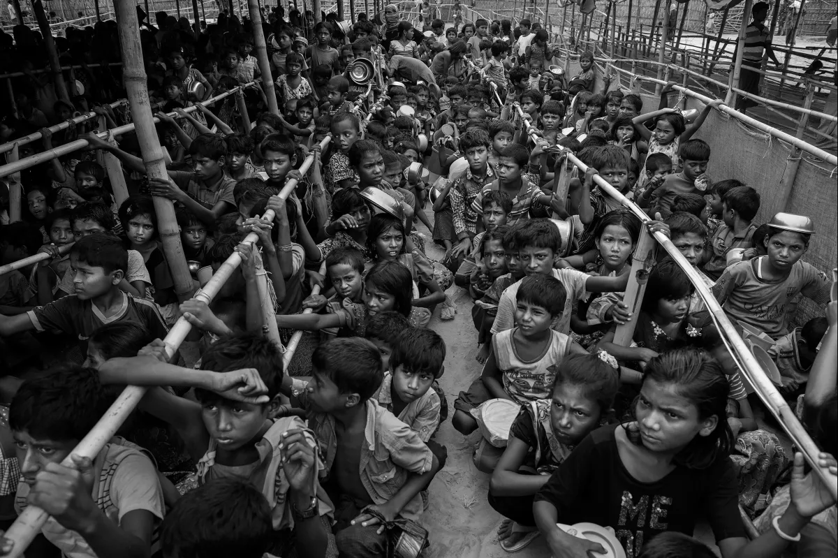 Des centaines d’enfants rohingyas, la plupart accroupis ou assis, font la queue pour de la nourriture. Les files d’attente sont séparées par de longues rangées de poteaux en bambou.