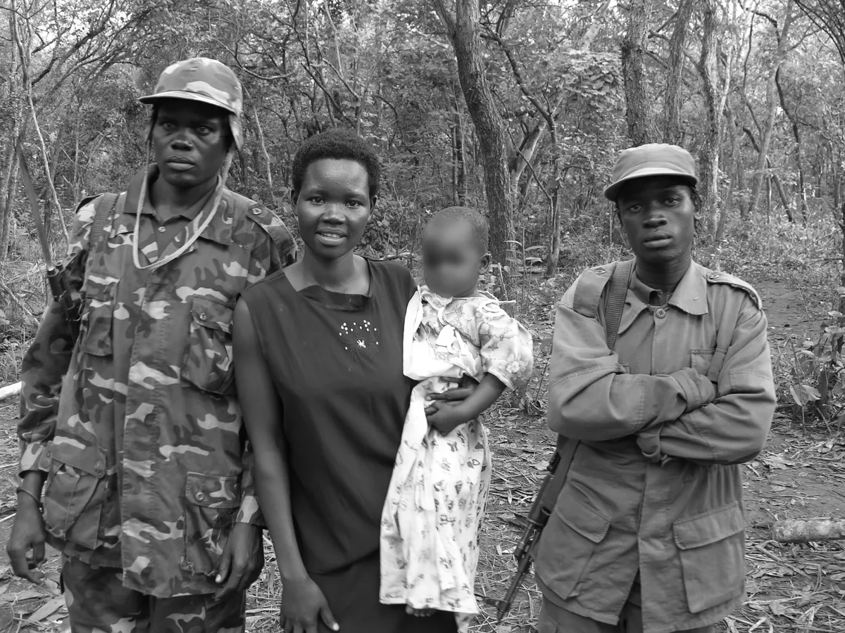 Une photo en noir et blanc d'une femme tenant un bébé dans ses bras, tandis que deux hommes en tenue militaire se tiennent à ses côtés. Ils sont debout devant une forêt et posent pour la photo.