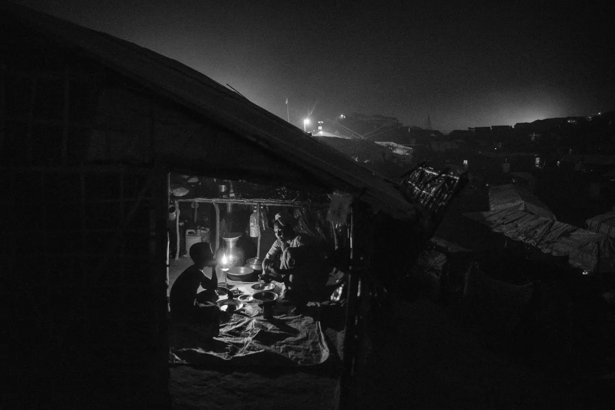 Une réfugiée rohingya sert de la nourriture à un garçon assis en face d’elle dans un abri sombre. L’intérieur est éclairé par une seule bougie.