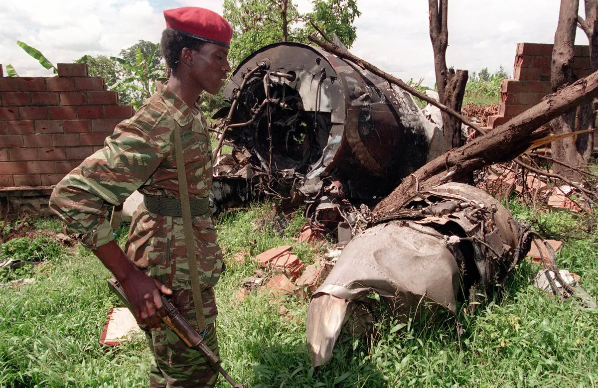 Un soldat portant un fusil, un béret rouge et un uniforme de camouflage se tient devant l’épave d’un avion au milieu des arbres.