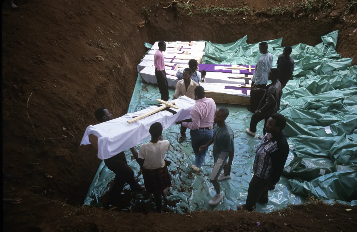 Plusieurs hommes portent un cercueil enveloppé de tissu et orné d’une grande croix en bois dans une grande fosse commune creusée dans la terre qui contient déjà plusieurs cercueils similaires.