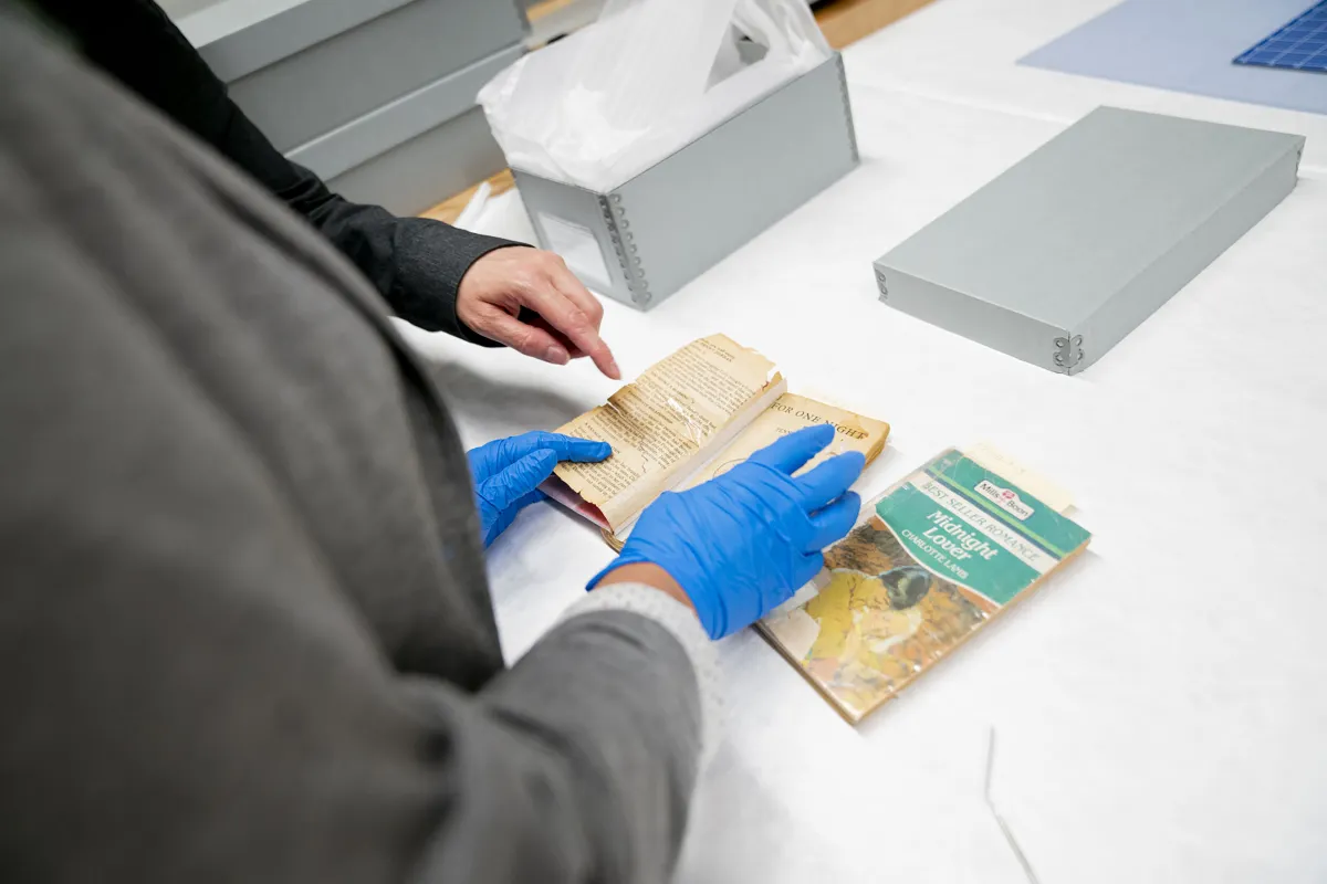 Des mains munies de gants de protection bleus tiennent un livre de poche ouvert à une page sur une table. La main d'une autre personne indique quelque chose dans le livre ouvert. Un autre livre est également posé sur la table, ainsi qu'une boîte et un couvercle en métal.